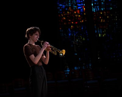 A women with short brown hair stands holding a trumpet with glowing stain glass behind her.