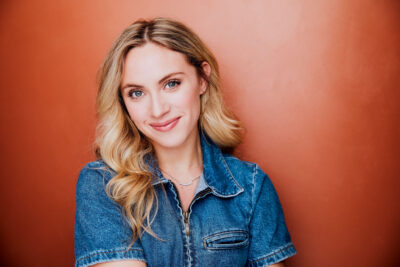 Girl with Blonde hair smiling wearing a blue denim shirt in front of a burnt orange background.